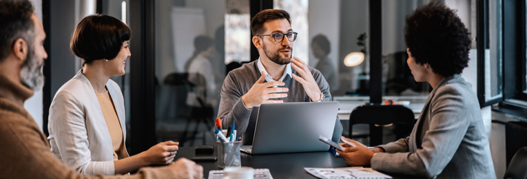 Four people in a boardroom meeting, engaged in discussion around a conference table.